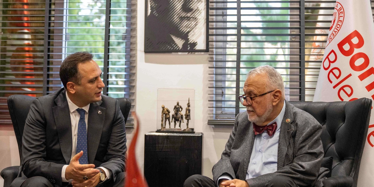Bornova Belediye Başkanı Ömer Eşmi, makamında bir konuğuyla oturmuş sohbet ediyor. Başkan koyu renk takım elbise ve mavi kravat giymiş, karşısında ise gri ceketli, gözlüklü ve papyonlu bir beyefendi oturuyor. Aralarındaki masanın üzerinde küçük bir asker figürü heykeli bulunuyor. Arka planda Bornova Belediyesi bayrağı ve duvarda Atatürk'ün silüetinin yer aldığı bir tablo görülüyor.