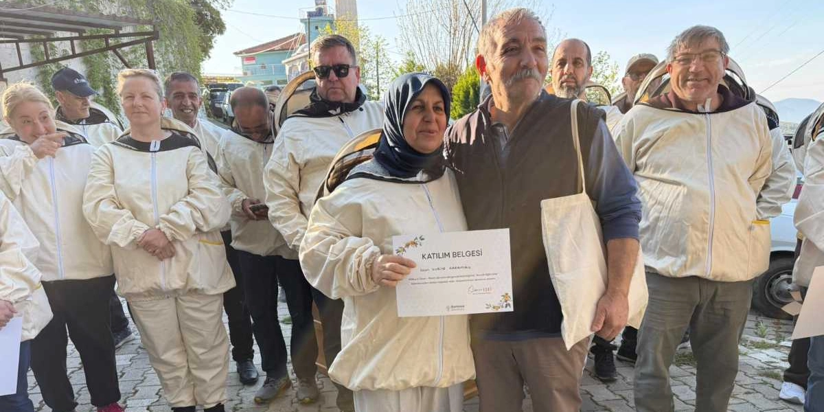 Arıcılık eğitimi almış bir grup kadın ve erkek, üzerlerinde koruyucu arıcı kıyafetleriyle açık havada yan yana duruyor. Grubun ön planında, başörtülü bir kadın elinde 