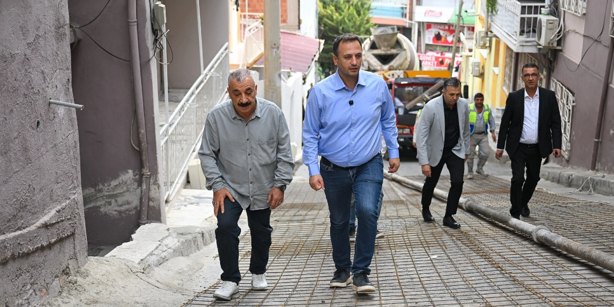 Bornova Belediye Başkanı Ömer Eşki ve beraberindeki heyet, beton dökümü için hazırlığı tamamlanmış, üzerinde çelik hasır döşeli olan yokuş bir sokakta incelemelerde bulunuyor. Arka planda bir beton mikseri ve çalışma yapan görevliler görülüyor.