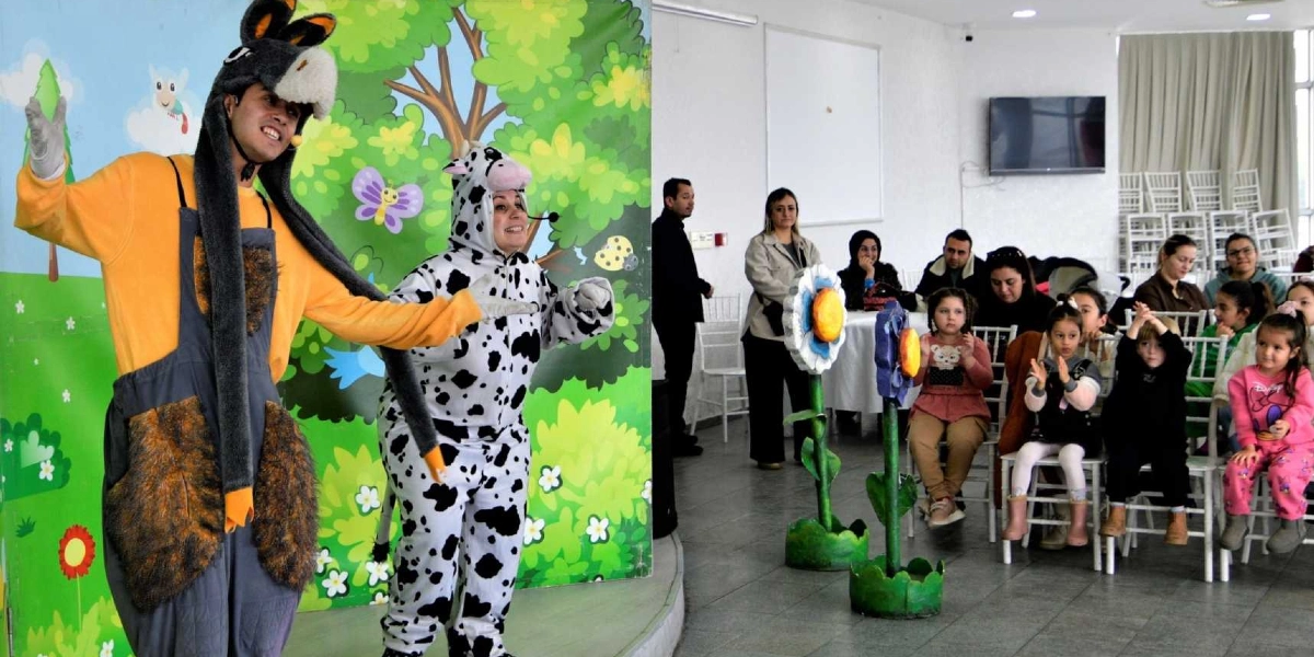 Sahne üzerinde eşek ve inek kostümü giymiş iki oyuncu çocuklara yönelik bir tiyatro gösterisi sergiliyor. Sahne dekorunda ağaçlar ve çiçekler yer alırken, izleyici kısmında oturan çocuklar ve yetişkinler gösteriyi ilgiyle takip ediyor.