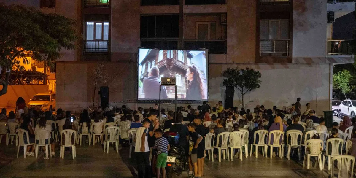 Gece vakti bir apartman bahçesinde kurulan dev ekranda film izleyen kalabalık bir grup insan. İnsanlar plastik sandalyelerde oturarak beyaz perdeye yansıtılan, eski bir ahşap evi ve iki kişiyi gösteren sahneyi izliyor. Çevrede ağaçlar, park halindeki araçlar ve apartman pencerelerinden yansıyan ışıklar görülüyor.