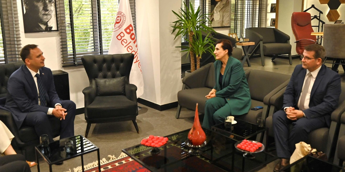 Modern bir ofis ortamında, Bornova Belediyesi bayrağının önünde karşılıklı koltuklarda oturan üç kişi bir toplantı gerçekleştiriyor. Solda lacivert takım elbiseli bir erkek, sağda ise yeşil ceketli bir kadın ve yanında gri takım elbiseli başka bir erkek yer alıyor. Önlerindeki cam sehpanın üzerinde kırmızı objeler ve içecekler bulunuyor.