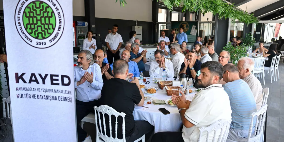 Karacaoğlan ve Yeşilova Mahalleleri Kültür ve Dayanışma Derneği'nin (KAYED) logosunun bulunduğu dikey bir pankartın önünde, bir restoranda toplanmış kalabalık bir grup insan görülüyor. Masalarda oturan katılımcılar alkışlıyor, bazıları ise birbirleriyle sohbet ediyor. Masaların üzerinde çay bardakları, tabaklar ve yiyecekler bulunuyor. Arka planda ayakta duran görevliler ve diğer masalarda oturan insanlar yer alıyor.