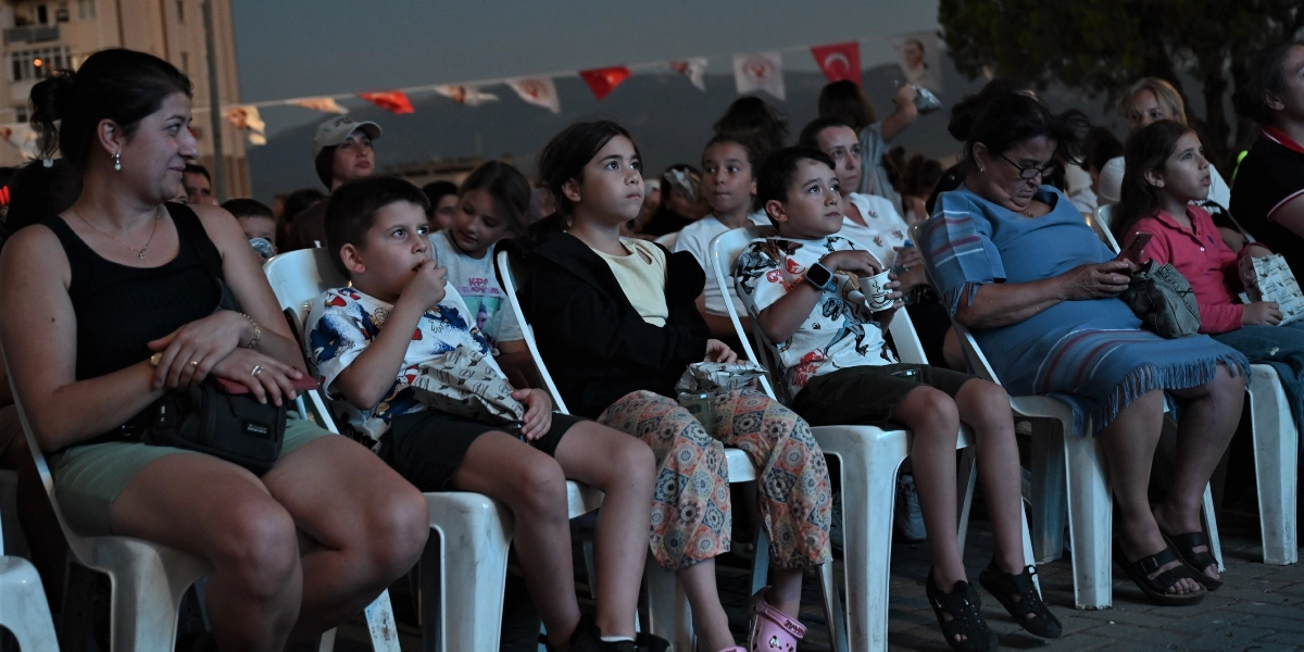 Açık havada, beyaz plastik sandalyelerde yan yana oturmuş, dikkatle bir şeyler izleyen bir grup çocuk ve yetişkin. Ön planda siyah atletli bir kadın, yanında elinde atıştırmalık paketiyle oturan bir erkek çocuk, onun yanında koyu renkli ceketli bir kız çocuğu, yanında beyaz desenli tişörtlü bir erkek çocuk ve en sağda mavi elbiseli, gözlüklü bir kadın yer alıyor. Arka planda ise Türk bayrakları ve süslemelerle donatılmış bir alan ile kalabalık bir izleyici grubu görülüyor.