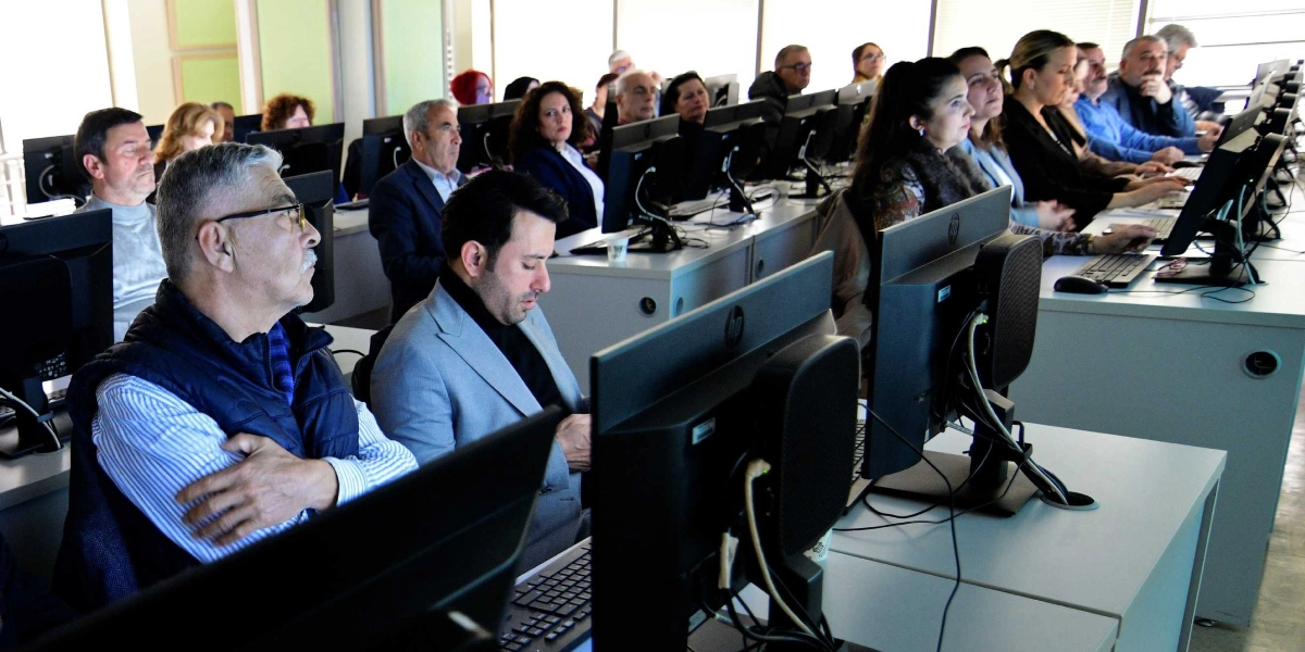 Bilgisayar laboratuvarı ortamında yan yana dizilmiş masalarda oturan, önlerindeki bilgisayar ekranlarına odaklanmış veya dinleme yapan çok sayıda kişi. Ön planda, çizgili gömlek ve lacivert yelek giyen gözlüklü bir erkek ile yanında gri ceketli bir başka erkek dikkat çekiyor. Katılımcıların çoğu iş kıyafetleri içinde, dikkatli bir şekilde eğitimi takip ediyor.