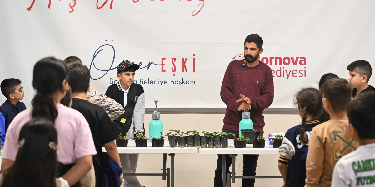 Sakallı bir eğitmen, bir grup çocuğa masa üzerindeki küçük saksılarda bulunan fidelerin bakımı hakkında uygulamalı bilgi veriyor. Eğitmen, elindeki pompalı su püskürtücüsünü kullanarak bitkileri nasıl sulayacaklarını gösterirken, çocuklar ilgiyle onu izliyor. Arka planda Bornova Belediyesi ve Belediye Başkanı Ömer Eşki'nin isminin yer aldığı beyaz bir afiş bulunuyor.