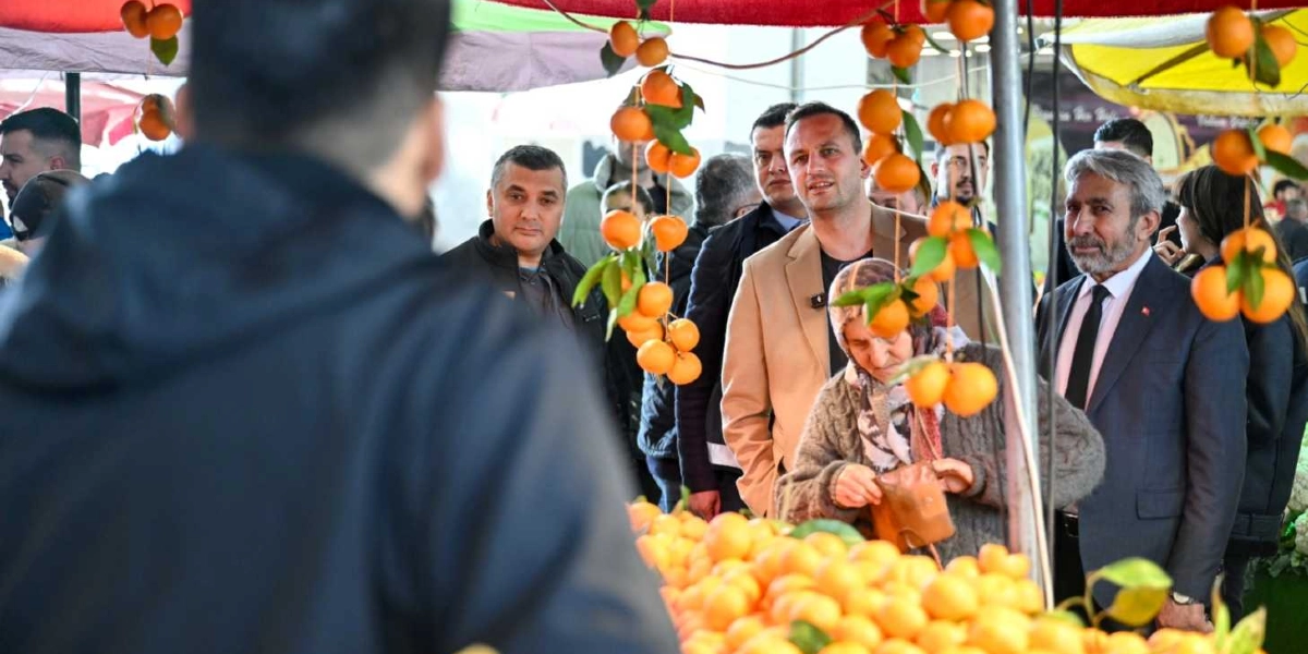 Bej ceketli bir erkek, yanında takım elbiseli ve kravatlı bir başka erkekle birlikte bir pazar tezgahını ziyaret ediyor. Tezgahın önünde alışveriş yapan başörtülü bir kadın ve çevrede onları izleyen diğer insanlar bulunuyor. Tezgahın üzerinde ve üst kısımdaki tenteye asılı şekilde bol miktarda taze mandalina sergileniyor.