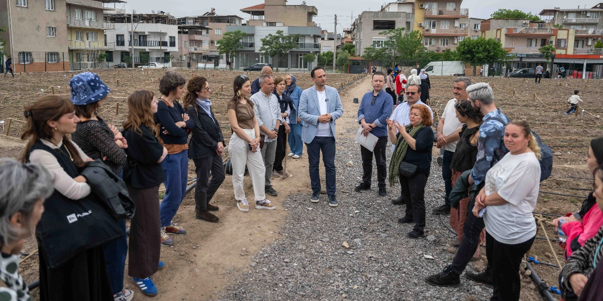 Bornova'da yeni oluşturulmuş bir kent bostanı alanında toplanmış, çoğunluğu kadınlardan oluşan bir grup insan, bir yetkilinin açıklamalarını dinliyor. Toprak zemin üzerinde, sulama boruları döşenmiş ve küçük işaret çubuklarıyla parsellere ayrılmış geniş bir alanda, arka planda apartmanların yer aldığı kentsel bir peyzaj görülüyor. Grup, çakıl taşlı bir yol üzerinde durmuş, dikkatle konuşmacıyı takip ediyor.