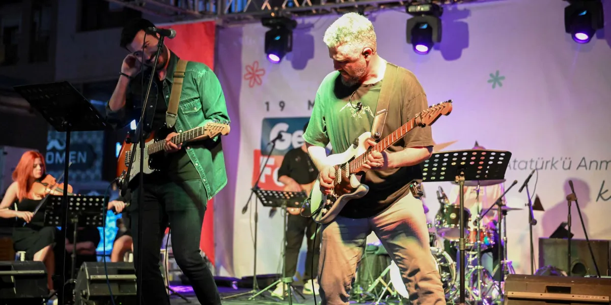 Açık havada kurulan bir sahnede canlı müzik performansı sergileyen bir grup müzisyen. Ön planda, yeşil gömlekli ve elektro gitar çalan bir erkek ile gri tişörtlü, beyaz elektro gitar çalan başka bir erkek yer alıyor. Sol tarafta keman çalan kızıl saçlı bir kadın, arka planda ise bateri çalan bir başka müzisyen görülüyor. Sahne arkasındaki panoda 
