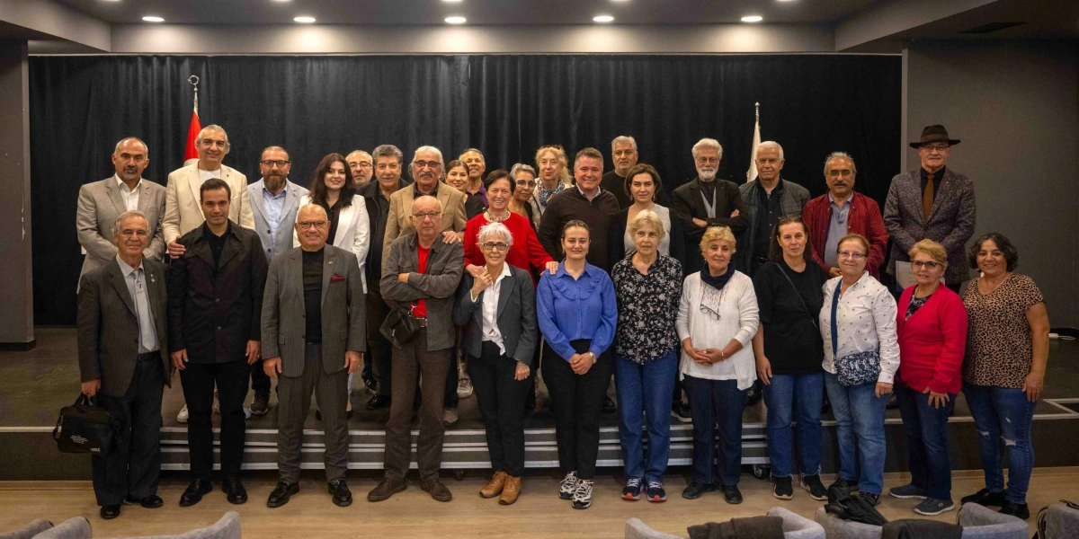 Kapalı bir sahnede, siyah bir perde önünde yan yana dizilmiş, farklı yaş ve giyim tarzlarına sahip yaklaşık otuz kişilik bir grup insan, kameraya gülümseyerek poz veriyor. Grubun arkasında, sahnenin sol tarafında bir Türk bayrağı yer alıyor. Katılımcılar arasında hem erkekler hem de kadınlar bulunuyor; birçoğu günlük kıyafetler, ceketler veya gömlekler giymiş durumda. Sahne aydınlatması tavan spotlarıyla sağlanıyor ve ön planda izleyici koltuklarının üst kısımları görünüyor.