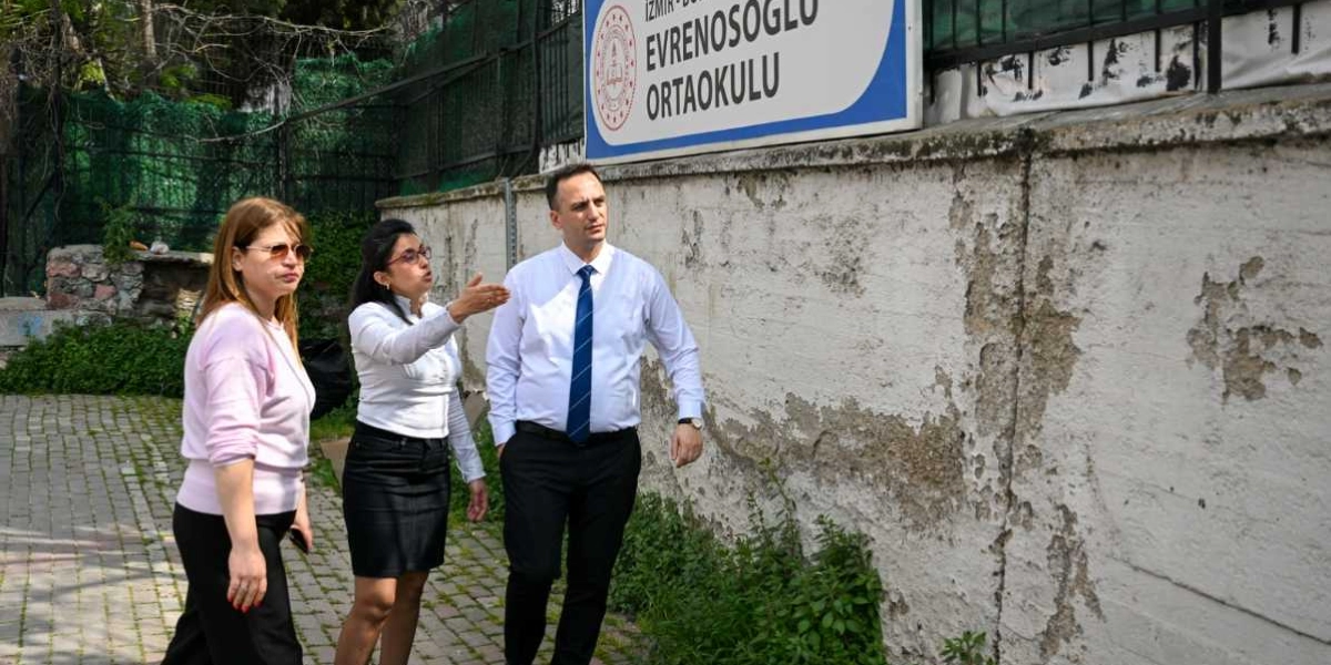 İzmir Evrenosoğlu Ortaokulu tabelasının önünde duran üç kişi, okulun dış duvarını inceleyerek sohbet ediyor. Beyaz gömlekli ve siyah etekli bir kadın eliyle duvarı işaret ederken, yanında duran beyaz gömlekli ve kravatlı bir erkek ile açık renk bluzlu bir kadın onu dinliyor. Okulun dış duvarı yer yer dökülmüş ve yıpranmış bir görünüme sahip.