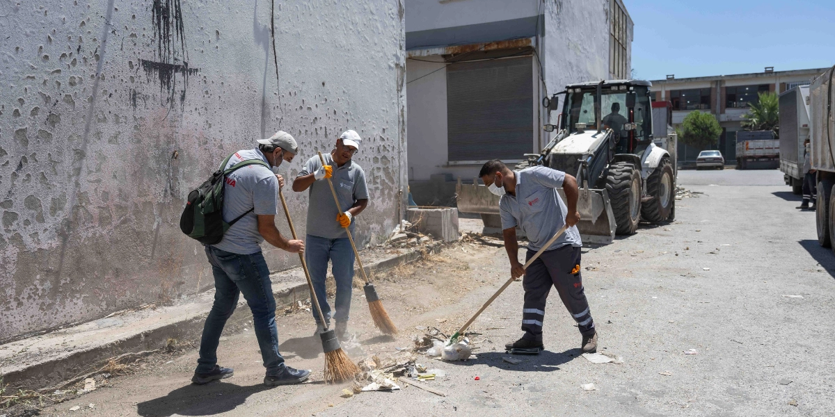 Bornova Belediyesi üniformalı üç çalışan, sanayi bölgesindeki bir sokakta süpürgelerle ayakkabı atıklarını ve çöpleri temizliyor. Çalışanların arkasında beyaz bir iş makinesi park halinde duruyor.