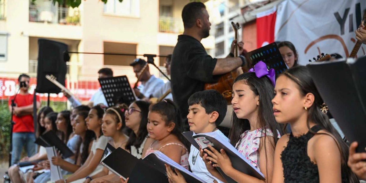 Açık havada düzenlenen bir etkinlikte, ellerinde nota sehpaları ve şarkı sözleri bulunan çocuklardan oluşan bir koro şarkı söylüyor. Ön planda, ellerinde dosyalarla şarkı söyleyen kız ve erkek çocukları yer alırken, arka planda onlara eşlik eden yetişkin bir müzisyen gitar çalıyor. Arka planda ise binalar ve etkinlik alanını çevreleyen bariyerler görülüyor.
