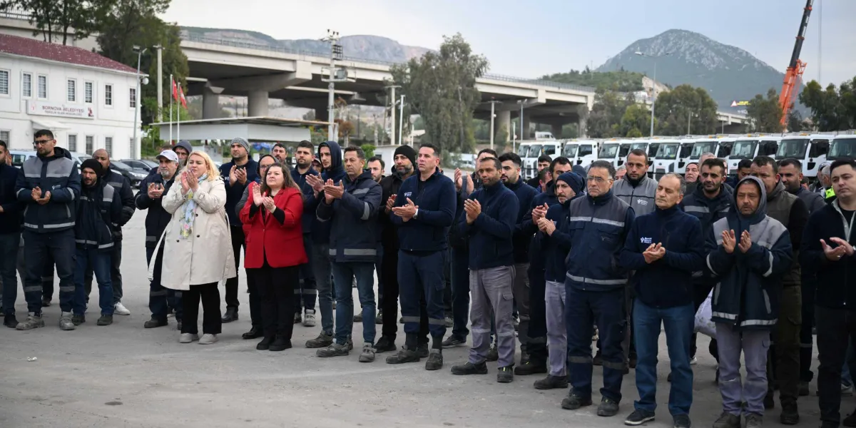 Bornova Belediyesi şantiye alanında, iş kıyafetleri giymiş kalabalık bir işçi grubu, Başkan Ömer Eşki'nin konuşmasını dinledikten sonra alkışlıyor. Arka planda Bornova Belediyesi Temizlik İşleri Müdürlüğü binası, park halindeki çok sayıda çöp kamyonu ve viyadükler yer alıyor.