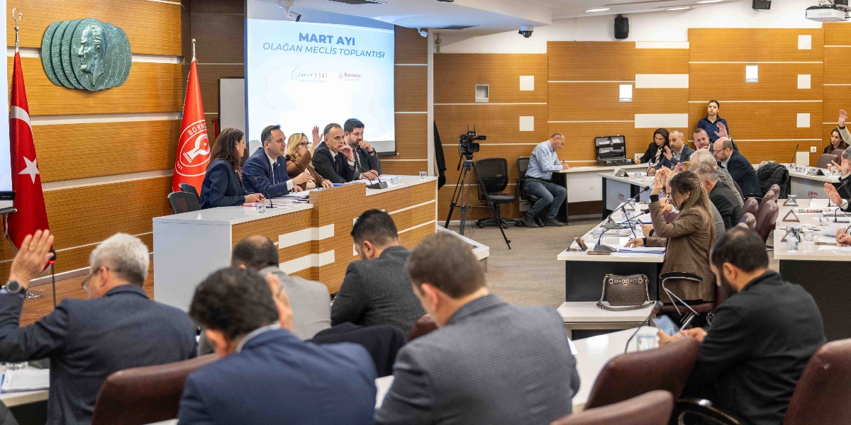 Bornova Belediyesi Mart ayı olağan meclis toplantısının gerçekleştirildiği salonda, katılımcılar masalarda otururken bazı üyeler oylama için ellerini kaldırmış durumda. Salonun ön kısmındaki kürsüde belediye yetkilileri yer alırken, arka plandaki ekranda toplantı başlığı yansıtılıyor. Salonun duvarında Atatürk rölyefi ile Türk bayrağı ve Bornova Belediyesi flaması bulunuyor.