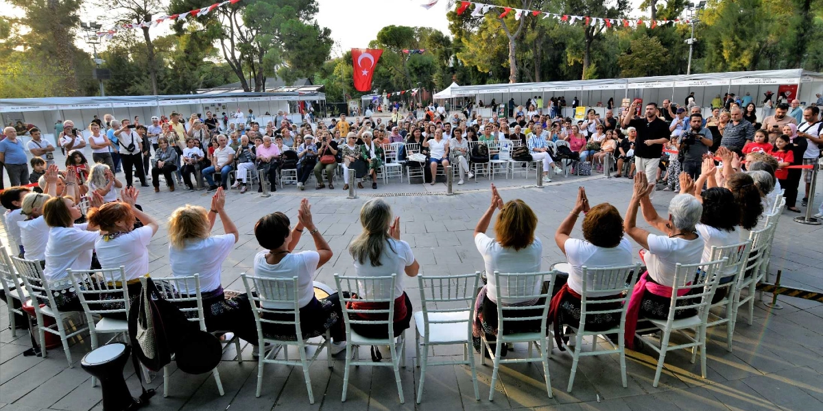 Açık havada düzenlenen bir kitap fuarı alanında, beyaz tişörtlü kadınlardan oluşan bir grup, sandalyelerde oturmuş elleriyle ritim tutarak performans sergiliyor. Grubun önünde, onları izleyen kalabalık bir seyirci kitlesi bulunuyor. Arka planda ise kitap stantları, ağaçlar ve asılı Türk bayrağı görülüyor.