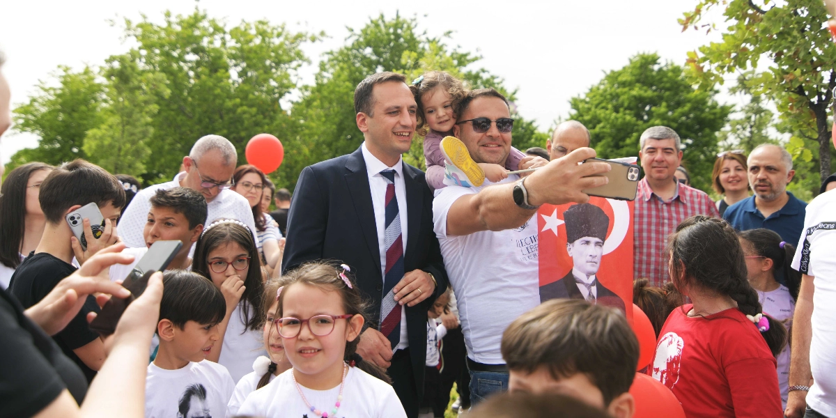 Açık havada, bir grup çocuk ve yetişkinin katıldığı 23 Nisan kutlamasında, beyaz tişörtlü bir adam omzunda küçük bir kız çocuğuyla birlikte kalabalıkla selfie çekiyor. Yanında lacivert takım elbiseli ve kravatlı bir erkek gülümsüyor. Çevredeki çocuklar ve yetişkinler neşeli bir şekilde poz verirken, ön planda üzerinde Atatürk fotoğrafı ve Türk bayrağı bulunan bir pankart dikkat çekiyor. Arka planda yeşil ağaçlar ve kırmızı balonlar görülüyor.