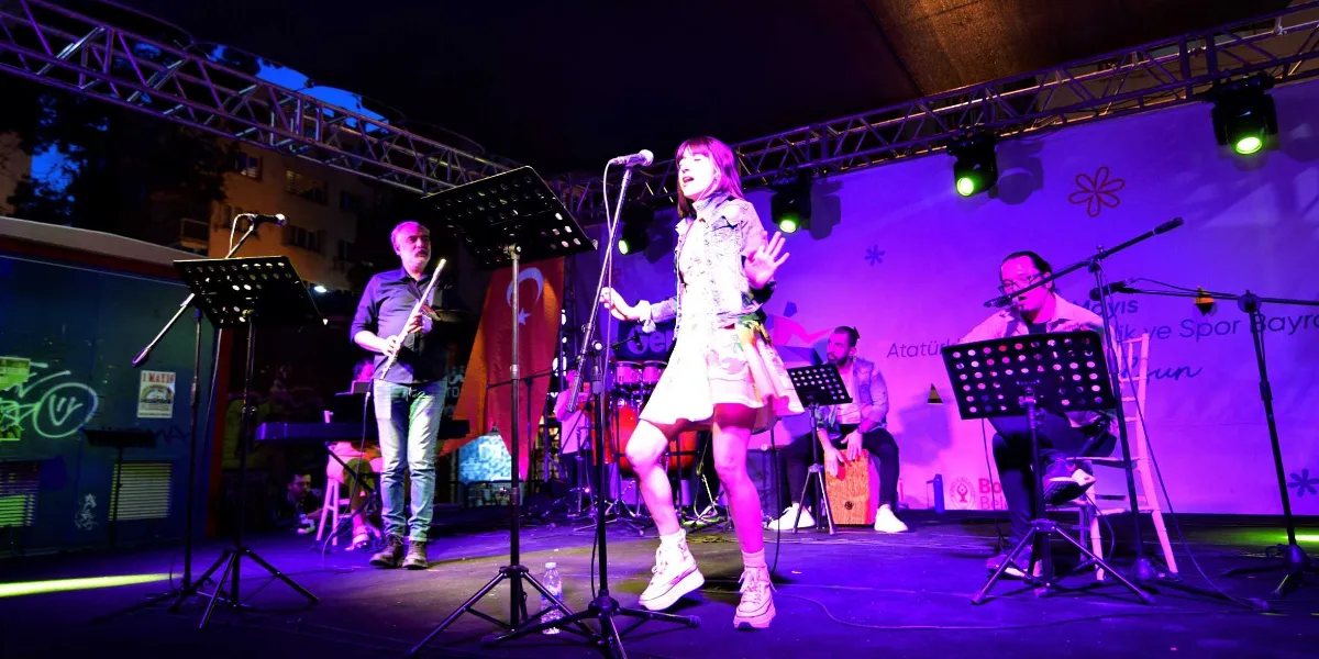 Bornova'da düzenlenen 19 Mayıs Atatürk'ü Anma, Gençlik ve Spor Bayramı kutlamaları kapsamında sahnede canlı müzik performansı sergileyen bir grup. Sahnenin merkezinde mikrofon başında şarkı söyleyen genç bir kadın solist, solunda flüt çalan bir müzisyen, sağında ise gitar çalan bir başka müzisyen yer alıyor. Arka planda ise perküsyon çalan bir kişi daha bulunuyor. Sahne mor ve mavi ışıklarla aydınlatılmış olup, arka planda Bornova Belediyesi logosunun ve 19 Mayıs kutlama mesajının yer aldığı bir afiş görülüyor.