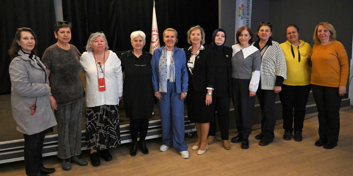 Kapalı bir mekanda, sahne üzerinde yan yana dizilmiş on kadın, gülümseyerek kameraya bakıyor. Kadınlar günlük ve şık kıyafetler içinde; aralarında ceket, hırka, kazak ve başörtüsü kullananlar bulunuyor. Arka planda siyah bir perde, Bornova Belediyesi'ne ait bir bayrak ve üzerinde 