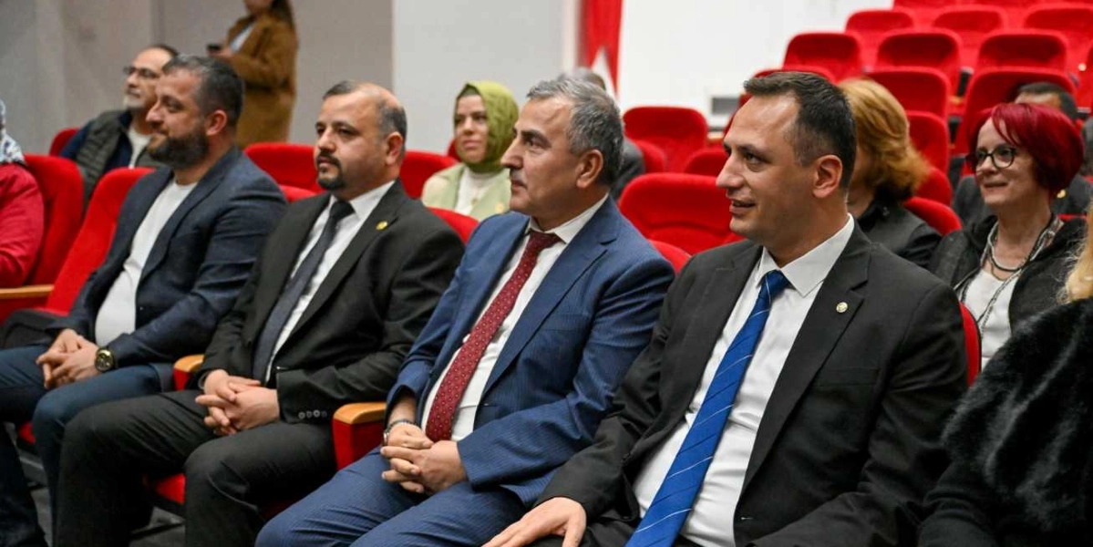 Konferans salonunda kırmızı koltuklarda yan yana oturan, takım elbiseli muhtarlar ve katılımcılar bir etkinliği izliyor. Ön sırada oturan dört erkekten üçü koyu renk takım elbise ve kravat takarken, en sağdaki kişi mavi kravatlı, yanındaki kişi ise bordo desenli kravatlıdır. Arka sıralarda başörtülü bir kadın ve kırmızı saçlı, gözlüklü bir kadın da dahil olmak üzere diğer katılımcılar dikkatle karşıya bakıyor.