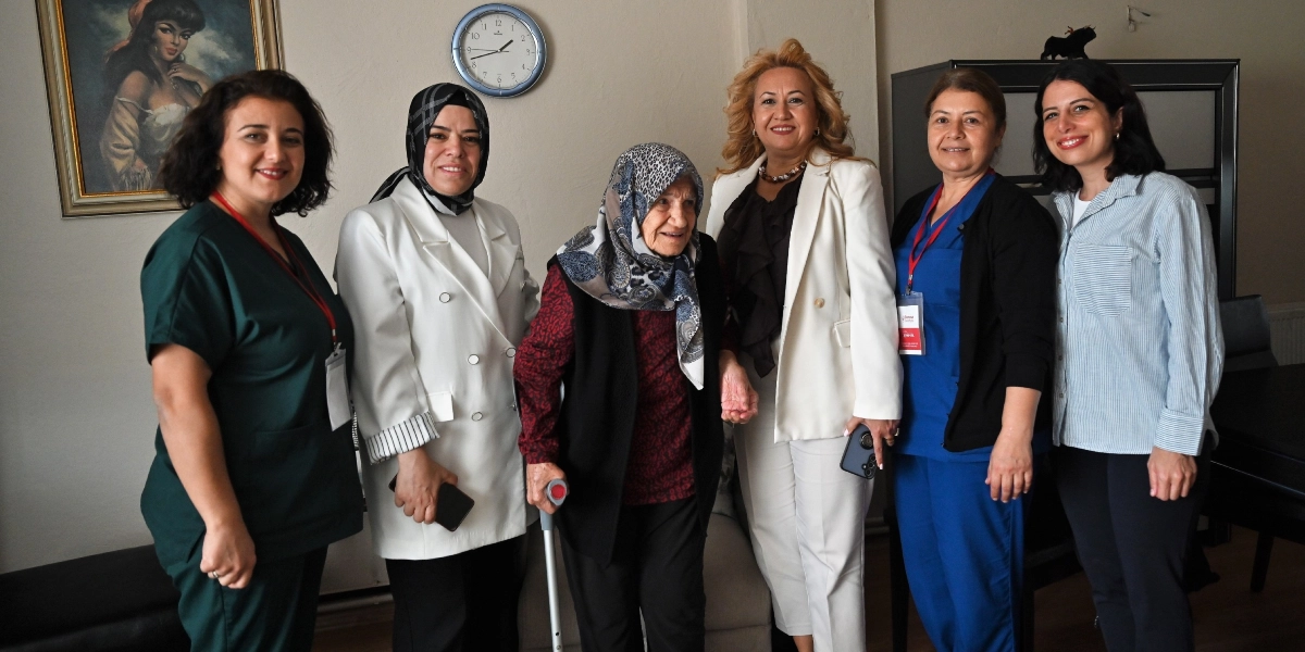 İç mekan bir odada, yaşlı bir kadını ziyaret eden beş kişilik bir grup kadın yan yana durarak gülümsüyor. Ortada duran yaşlı kadın, başında desenli bir eşarp, üzerinde bordo bir üst ve siyah bir yelek ile koltuk değneği kullanarak ayakta duruyor. Yanındaki grupta, beyaz ceketli, lacivert üniformalı ve günlük kıyafetli kadınlar yer alıyor. Arka planda duvarda bir saat ve çerçeveli bir tablo bulunuyor.
