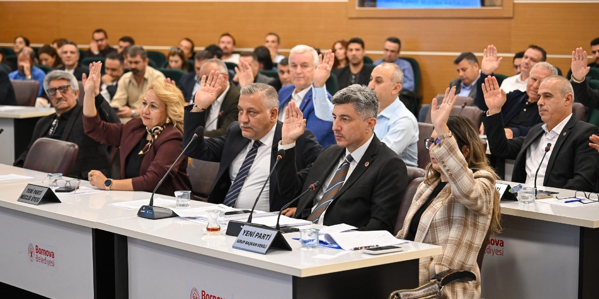 Bornova Belediyesi meclis salonunda, masalarda oturan çok sayıda meclis üyesinin bir oylama sırasında ellerini kaldırarak oy kullandığı toplantı anı. Ön planda, üzerinde 
