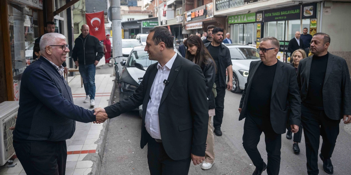 Siyah takım elbiseli bir erkek, Çamdibi'ndeki bir sokakta yaşlı bir vatandaşla el sıkışıyor. Başkanın etrafında korumaları ve çalışma arkadaşları yer alırken, arka planda esnafların bulunduğu hareketli bir sokak ve asılı bir Türk bayrağı görülüyor.