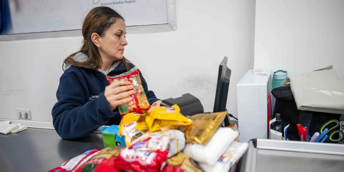 Üzerinde Bornova Belediyesi logosu bulunan lacivert bir polar giymiş kadın çalışan, bir market kasasında çeşitli gıda ürünlerini barkod okuyucudan geçirerek paketliyor.
