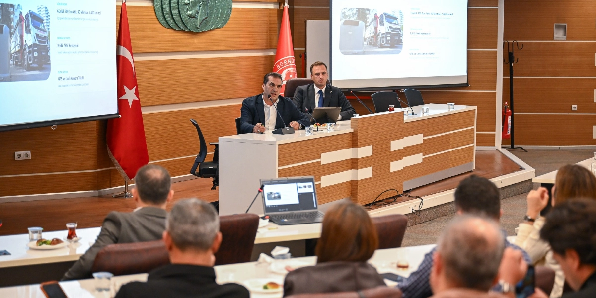 Bornova Belediyesi meclis salonunda, kürsüde konuşma yapan bir yetkili ve yanında oturan bir yönetici, katılımcılara sunum yapıyor. Arkadaki projeksiyon ekranında Bornova'daki temizlik hizmetlerine dair veriler yer alıyor; günlük 700 ton atık toplandığı, 9 bin 643 aktif konteyner bulunduğu ve araçların GPS ile canlı kamera sistemleriyle takip edildiği bilgileri paylaşılıyor. Salonun ön tarafında dinleyiciler masalarda oturuyor, arka planda ise Türk bayrağı ve Bornova Belediyesi'nin logosu bulunuyor.