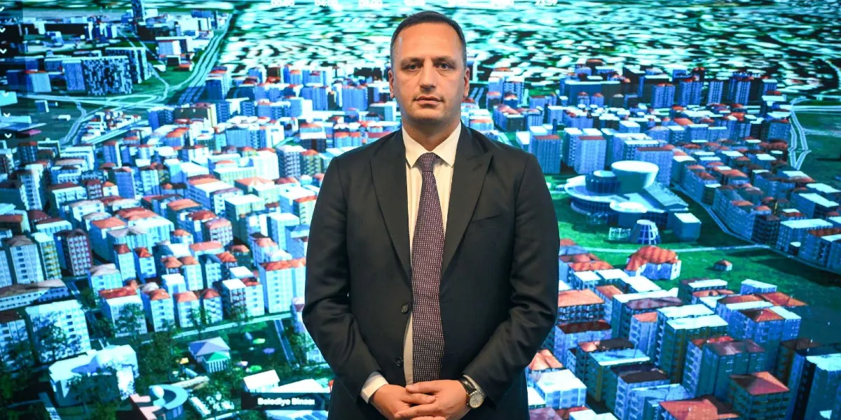 Koyu renk takım elbiseli ve kravatlı bir erkek, arkasında Bornova'nın dijital ikizini temsil eden üç boyutlu şehir modellemesinin yer aldığı dev bir ekranın önünde duruyor.