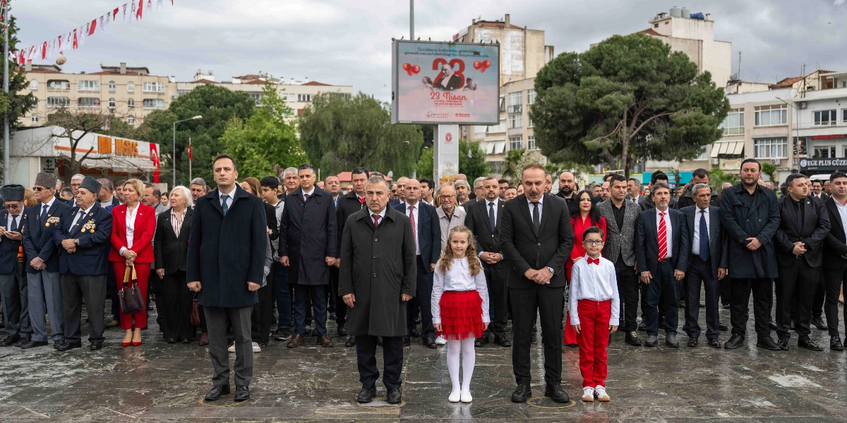 Açık havada, resmi tören alanında yan yana dizilmiş kalabalık bir grup insan, saygı duruşunda bulunuyor. Ön sırada, takım elbiseli yetişkinlerin arasında, beyaz gömlekli ve kırmızı etekli bir kız çocuğu ile beyaz gömlekli, kırmızı papyonlu ve kırmızı pantolonlu bir erkek çocuğu yer alıyor. Arka planda, üzerinde Atatürk görseli ve 