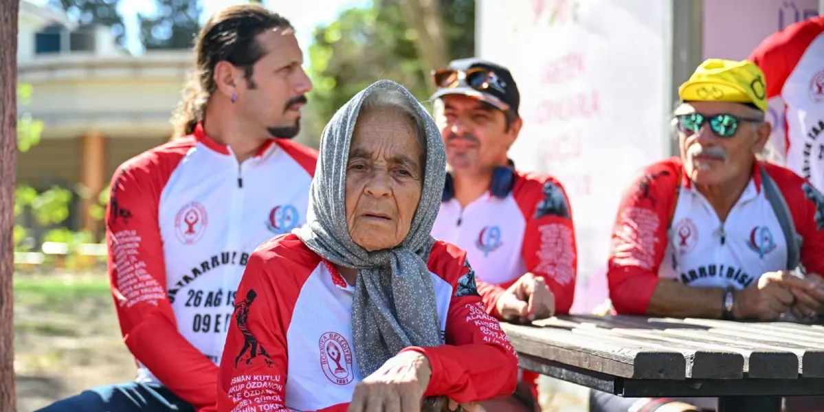 Başörtülü yaşlı bir kadın, arkasında Bornova Belediyesi logolu, kırmızı beyaz bisikletçi formaları giymiş üç erkekle birlikte dış mekanda bir masada oturuyor. Kadın doğrudan kameraya bakarken, arkasındaki erkekler farklı yönlere bakıyor; en sağdaki erkek sarı bir bisikletçi şapkası ve güneş gözlüğü takıyor. Formaların üzerinde 