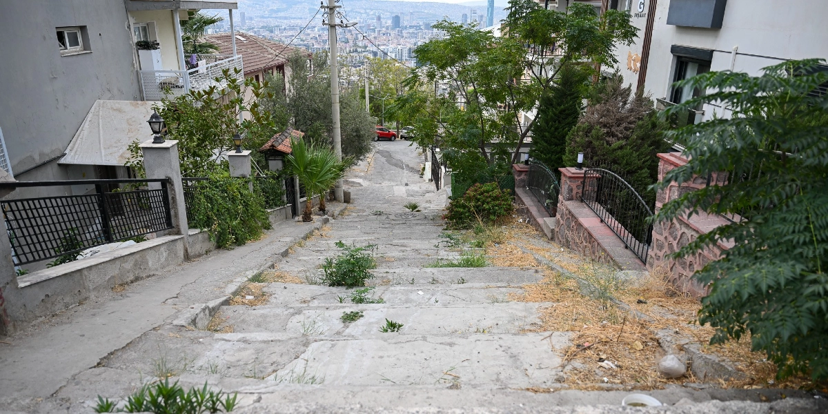 Bornova'da yokuş aşağı uzanan, yer yer otların çıktığı beton basamaklı bir merdiven yolu. Merdivenlerin iki yanında binalar, bahçe duvarları, ağaçlar ve demir korkuluklar bulunuyor. Merdivenlerin bittiği noktada asfalt bir yol ve arka planda geniş bir şehir manzarası görülüyor.