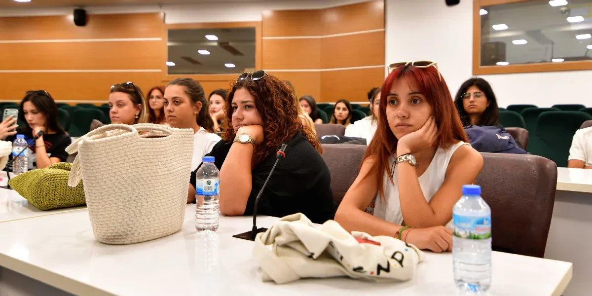 Konferans salonunda sıralarda oturan ve dikkatle dinleyen Peyzaj Mimarlığı öğrencileri. Ön planda, masaların üzerinde su şişeleri, bez çantalar ve kişisel eşyalar bulunuyor; öğrenciler ise masalara yaslanmış veya elleri çenelerinde sunumu takip ediyor.