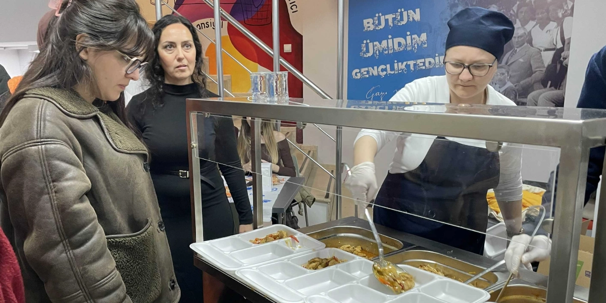 Bir yemekhanede, beyaz önlüklü ve lacivert kep takan bir görevli, paslanmaz çelik bir tezgahın arkasında plastik bölmeli tabaklara yemek servisi yapıyor. Görevlinin yanında, sırasını bekleyen iki kadın müşteri ilgiyle yemekleri izliyor. Arka plandaki duvarda, Mustafa Kemal Atatürk'ün fotoğrafının üzerinde 