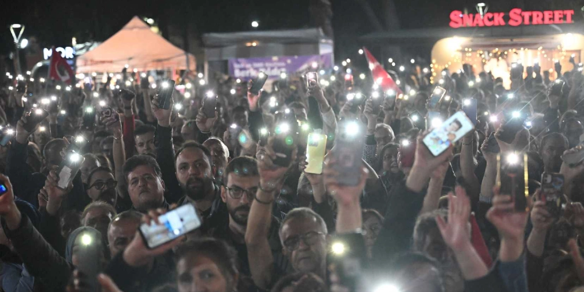 Gece vakti açık havada düzenlenen bir etkinlikte, kalabalık bir grup ellerindeki cep telefonlarının ışıklarını havaya kaldırarak coşkuyla kutlama yapıyor. Arka planda 