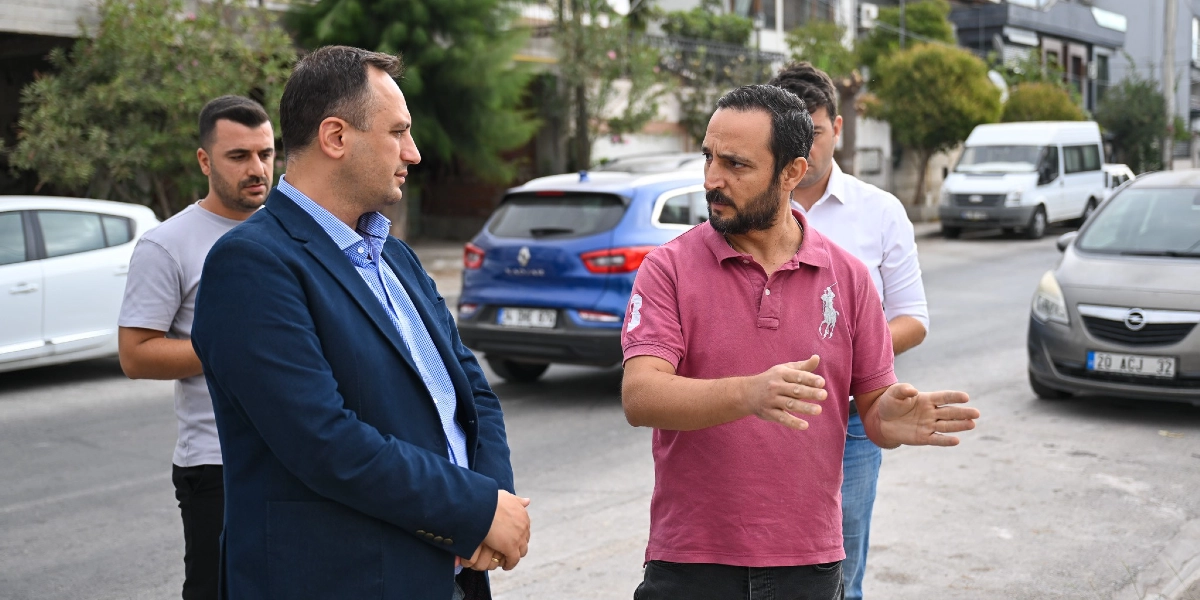 Sokakta bir arada duran dört erkek, bir konu hakkında konuşuyor. Ön planda, lacivert ceketli ve mavi gömlekli bir adam, yanında duran ve elleriyle tarifte bulunan pembe polo yaka tişörtlü adamı dinliyor. Arkalarında ise gri bir tişört giyen başka bir kişi ve beyaz bir gömlekli bir adam daha yer alıyor. Arka planda park halindeki araçlar ve sokak manzarası görülüyor.