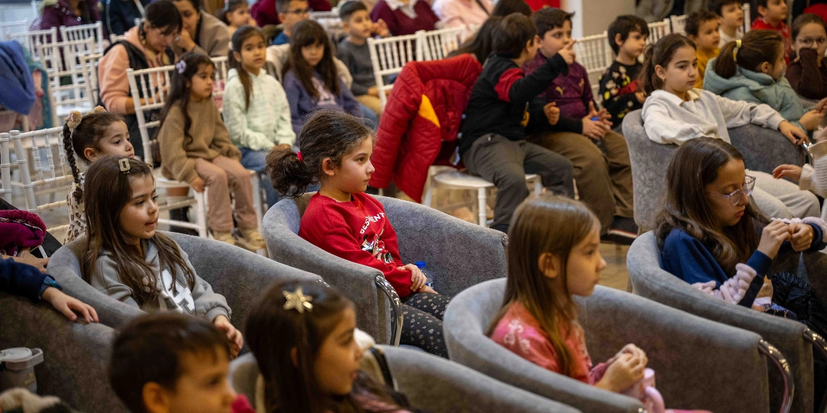 Bir tiyatro salonunda veya etkinlik alanında, gri koltuklarda yan yana oturmuş, dikkatle sahneye bakan çok sayıda çocuk ve onlara eşlik eden yetişkinler. Çocukların bir kısmı kışlık kıyafetler içinde, bazıları ise heyecanla çevrelerini inceliyor.