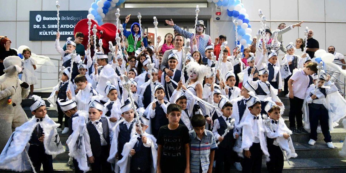 Bornova Belediyesi Dr. S. Sırrı Aydoğan Kültür Merkezi önündeki merdivenlerde, sünnet kıyafetleri giymiş ve ellerinde asalar tutan çok sayıda çocuk neşeyle poz veriyor. Çocukların arasında palyaço kostümlü animatörler ve yetişkinler yer alıyor; giriş kapısı ise mavi ve beyaz balonlarla süslenmiş durumda.
