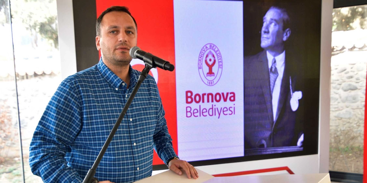 Mavi kareli gömlek giymiş bir erkek, önündeki mikrofona konuşma yapıyor. Arkasındaki ekranda Bornova Belediyesi logosu ve Mustafa Kemal Atatürk'ün siyah beyaz bir fotoğrafı yer alıyor.