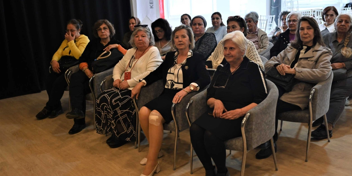 Kapalı bir salonda, gri koltuklarda yan yana oturmuş, dikkatle dinleyen çeşitli yaş gruplarından kadınlar grubu. Ön sırada, siyah beyaz desenli bluz ve ceket giymiş, dizinde sargı bulunan bir kadın ile yanında siyah kıyafetli başka bir kadın yer alıyor. Arka sıralarda ise farklı kıyafetler içinde, toplantıyı takip eden diğer kadın katılımcılar bulunuyor.