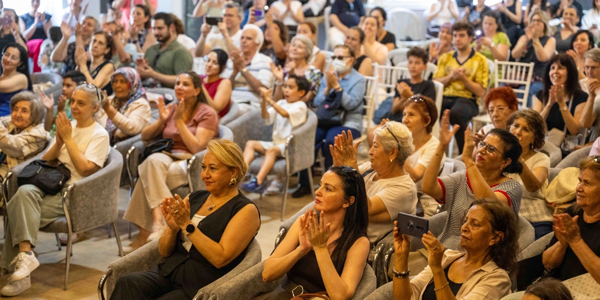 Kapalı bir salonda, gri koltuklarda oturan ve büyük bir çoğunluğu orta yaş ve üzeri kadınlardan oluşan kalabalık bir izleyici grubu, bir mezuniyet törenini veya etkinliği alkışlıyor. Ön planda, şık giyimli kadınlar dikkatle izleyip alkışlarken, bazıları cep telefonlarıyla fotoğraf çekiyor. Arka sıralarda ise farklı yaş gruplarından insanlar, heyecanlı ve mutlu bir ifadeyle töreni takip ediyor.