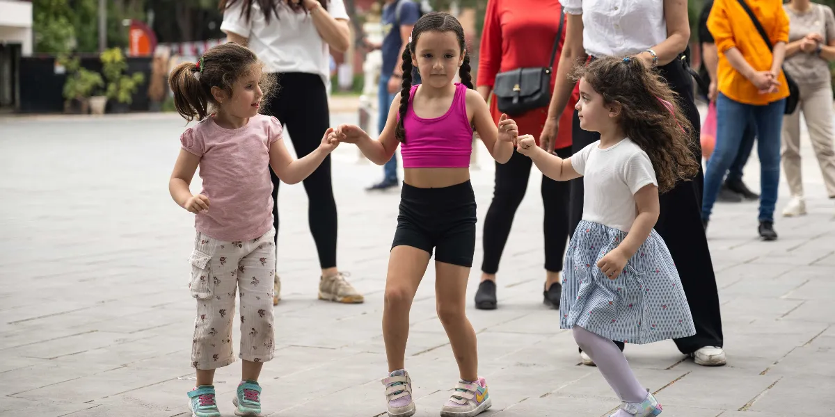 Açık havada el ele tutuşarak neşeyle dans eden üç küçük kız çocuğu. Soldaki kız pembe tişört ve desenli pantolon, ortadaki kız fuşya spor üst ve siyah şort, sağdaki kız ise beyaz tişört ve mavi beyaz çizgili etek giyiyor. Çocukların arkasında, onları izleyen yetişkinlerin bacakları ve vücutlarının bir kısmı görünüyor.