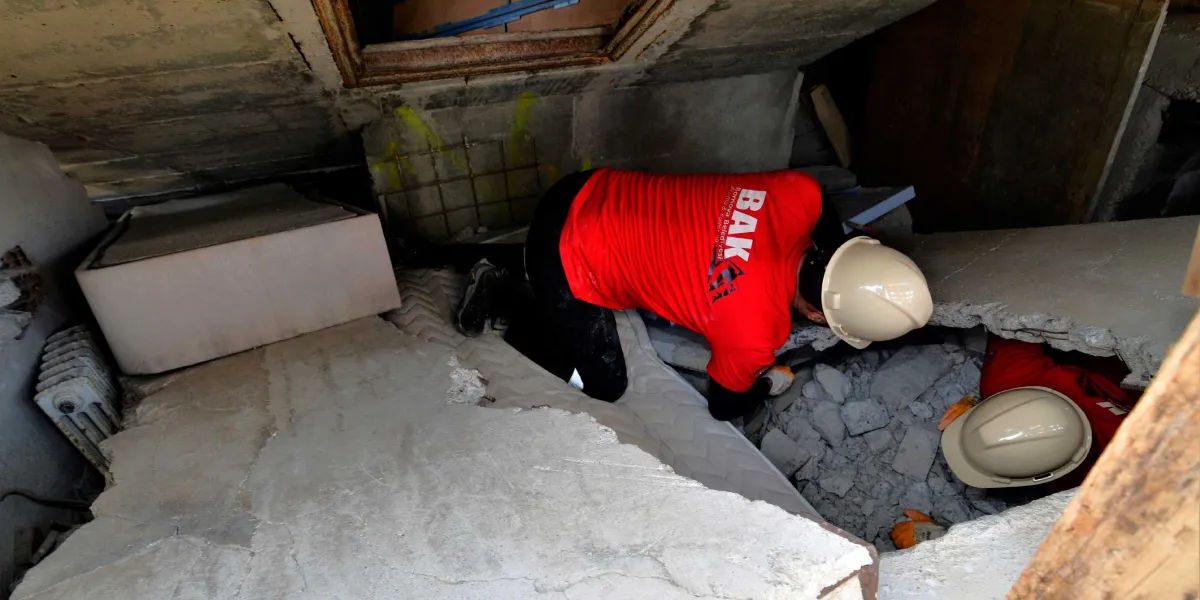 Üzerinde Bornova Belediyesi Arama Kurtarma (BAK) yazılı kırmızı tişörtler ve bej rengi baretler giyen iki arama kurtarma görevlisi, yıkılmış bir binanın enkazı arasında, beton blokların altında çalışma yürütüyor.