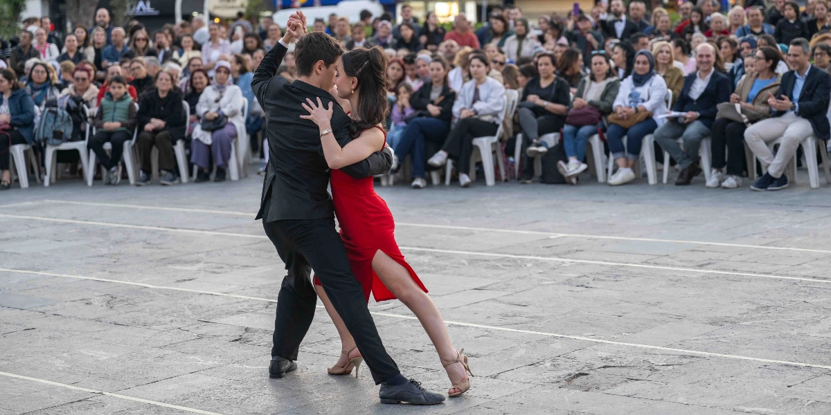 Açık havada, taş zeminli bir meydanda siyah takım elbiseli bir erkek ile kırmızı askılı elbise giyen bir kadının tango yaptığı an. Çift birbirine yakın durarak dans ederken, arkalarında beyaz sandalyelere oturmuş ve onları ilgiyle izleyen kalabalık bir seyirci grubu bulunuyor.