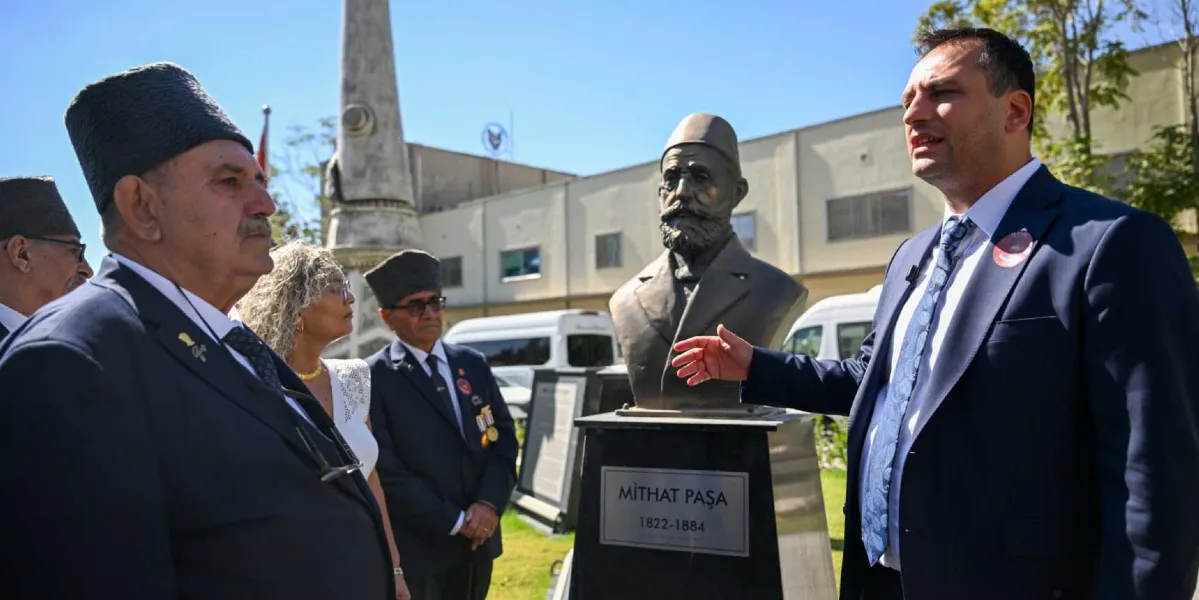Açık havada, Mithat Paşa'nın büstünün önünde duran bir grup insan görülüyor. Sağ tarafta lacivert takım elbiseli bir erkek, el hareketleriyle konuşma yapıyor. Karşısında ise geleneksel kalpak giymiş gaziler ve bir kadın yer alıyor. Büstün kaidesinde 
