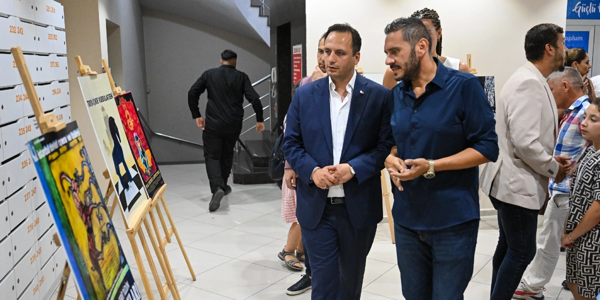 Modern bir binanın iç mekanında, şövale üzerinde sergilenen sanat afişlerini inceleyen bir grup insan. Ön planda, lacivert takım elbiseli bir erkek ile yanındaki koyu mavi gömlekli bir erkek, afişlere bakarak sohbet ediyor. Arka planda, üzerinde numaralandırılmış posta kutularına benzer bölmelerin bulunduğu beyaz bir duvar ve merdivenlere doğru yürüyen siyah giyimli bir kişi yer alıyor. Sağ tarafta ise sergiyi gezen diğer ziyaretçiler ve çocuklar görülüyor.
