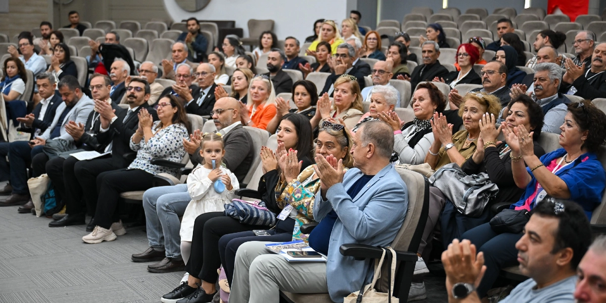 Konferans salonunda oturan ve alkışlayan kalabalık bir izleyici grubu. Ön sıralarda beyaz elbiseli küçük bir kız çocuğu ve yanında oturan yetişkinler dikkat çekerken, salonun genelinde farklı yaş gruplarından insanlar sahneye doğru dönmüş şekilde oturuyor.