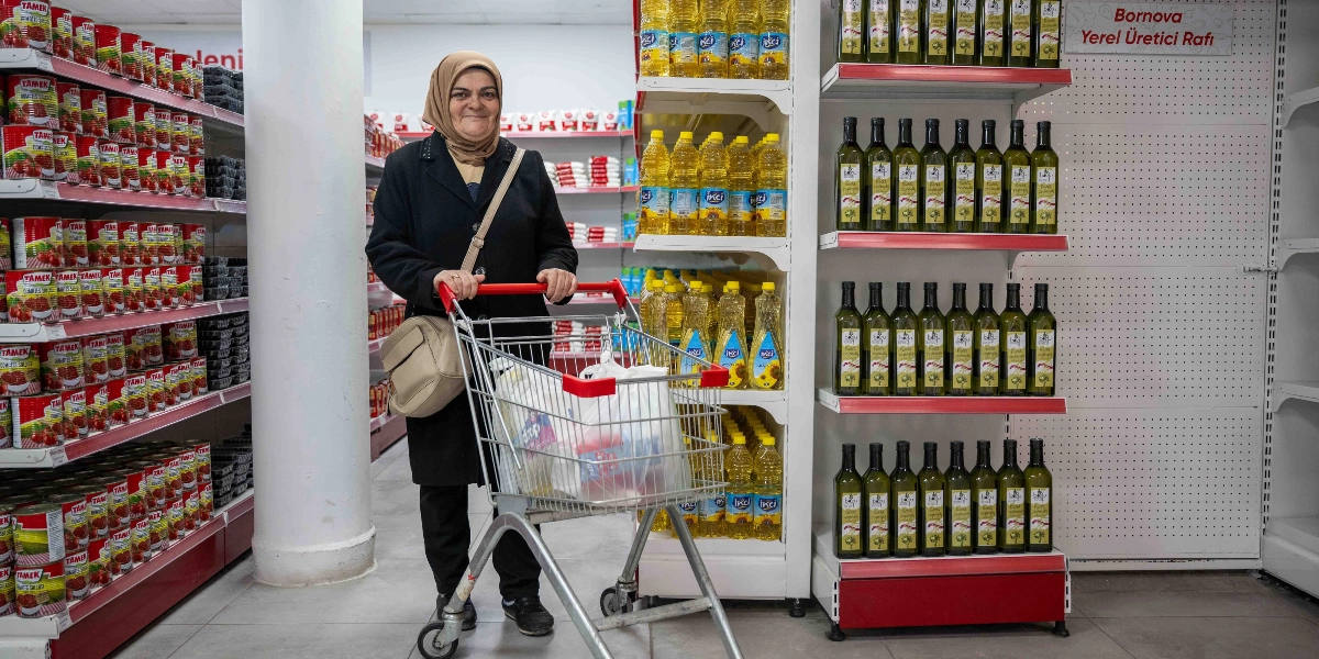 Başörtülü bir kadın, market reyonları arasında içinde alışveriş poşetleri bulunan bir market arabasını sürerken gülümseyerek kameraya bakıyor. Kadının solunda konserve ürünlerle dolu raflar, sağında ise üzerinde 