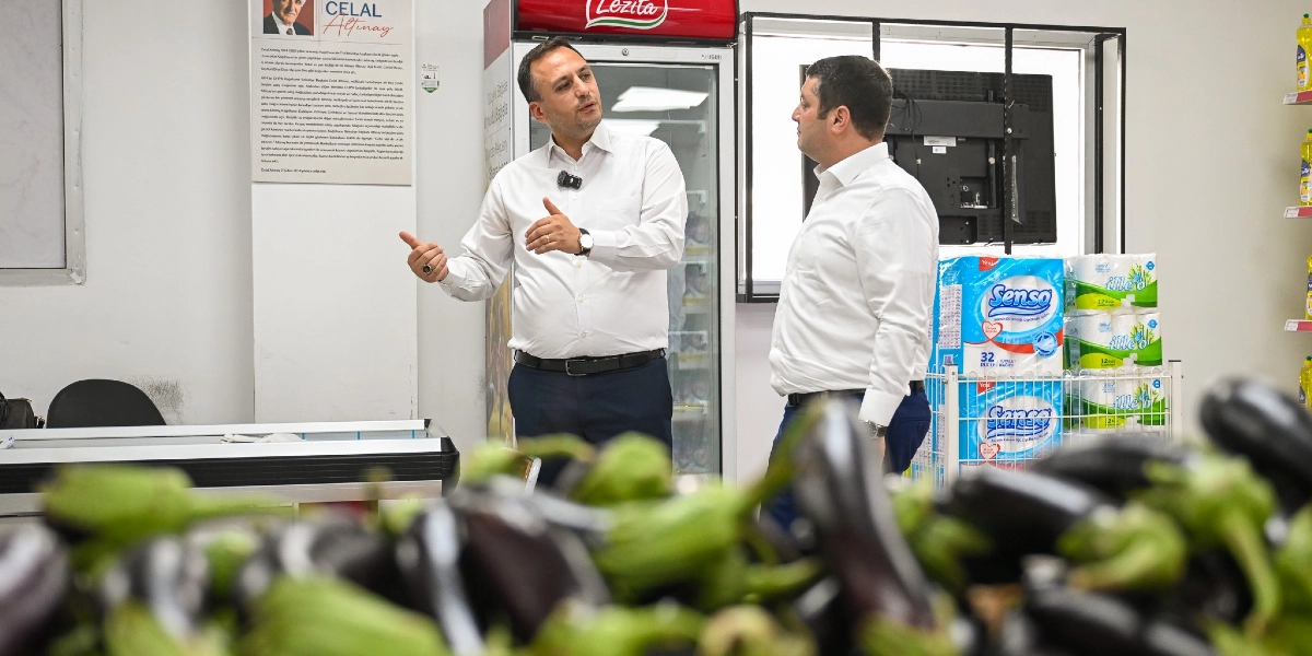 Beyaz gömlekli iki erkek, bir marketin içinde birbirleriyle konuşuyor. Soldaki kişi el hareketleriyle bir şeyler anlatırken, sağdaki kişi onu dinliyor. Arka planda bir buzdolabı, raflarda tuvalet kağıtları ve duvarda Celal Altınay'a ait bir bilgilendirme metni bulunuyor. Ön planda ise net olmayan bir şekilde patlıcanlar görülüyor.